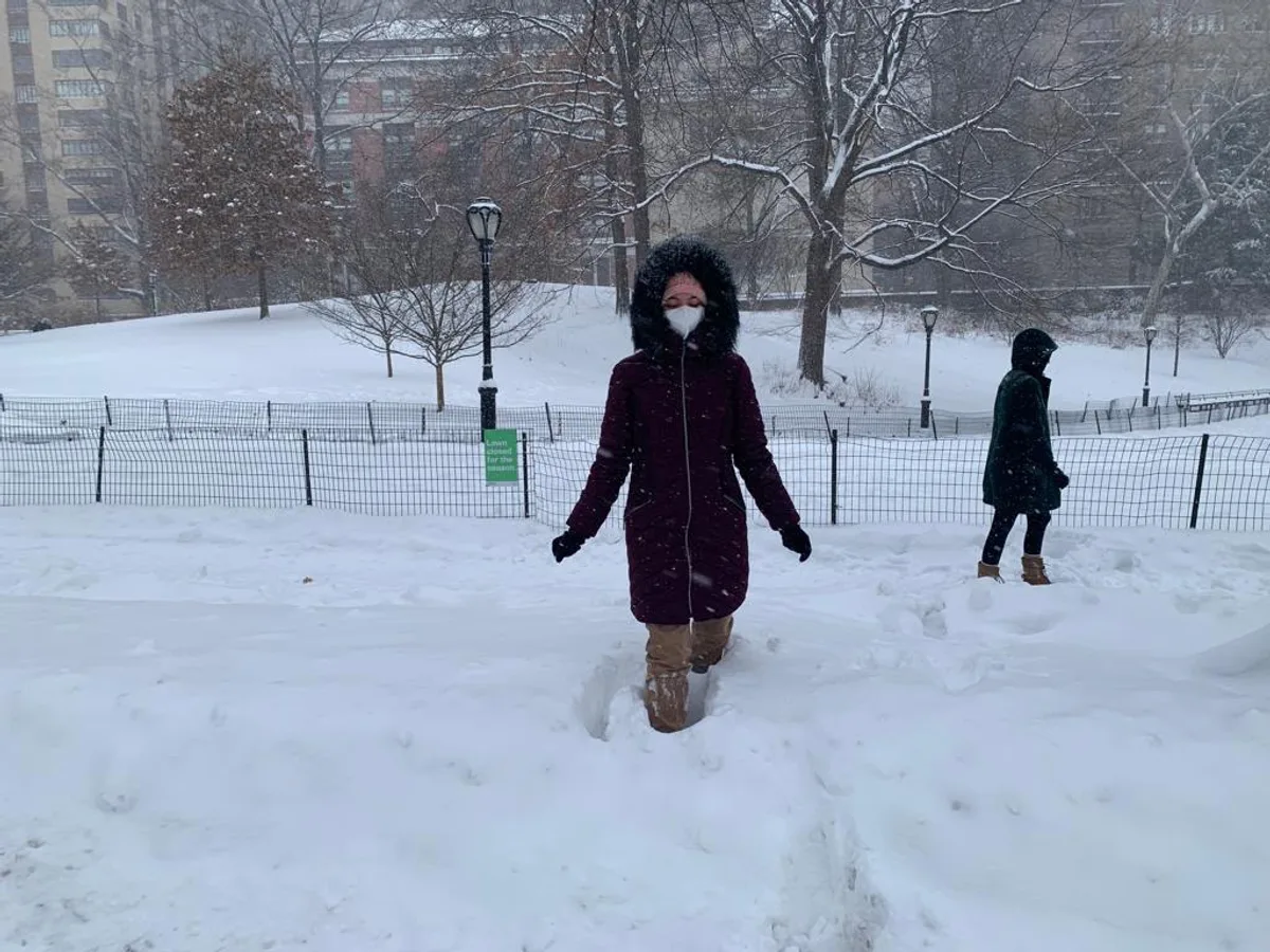 Julia de casaco pesado em pé na neve, num parque com prédios ao fundo