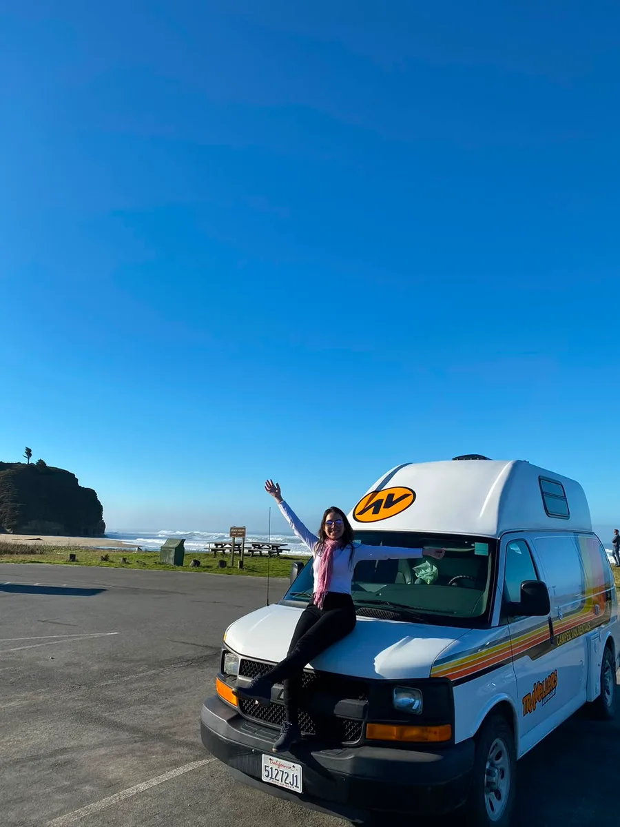 Julia sorrindo em pé sobre a frente de uma van de viagem, com o braço erguido, à beira da estrada