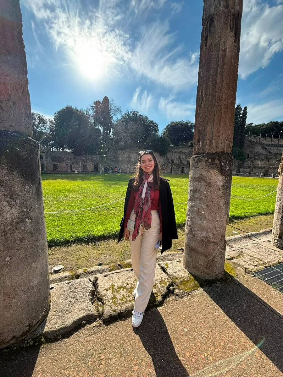 Julia sorrindo entre duas colunas antigas de um sítio arqueológico romano, num dia de sol